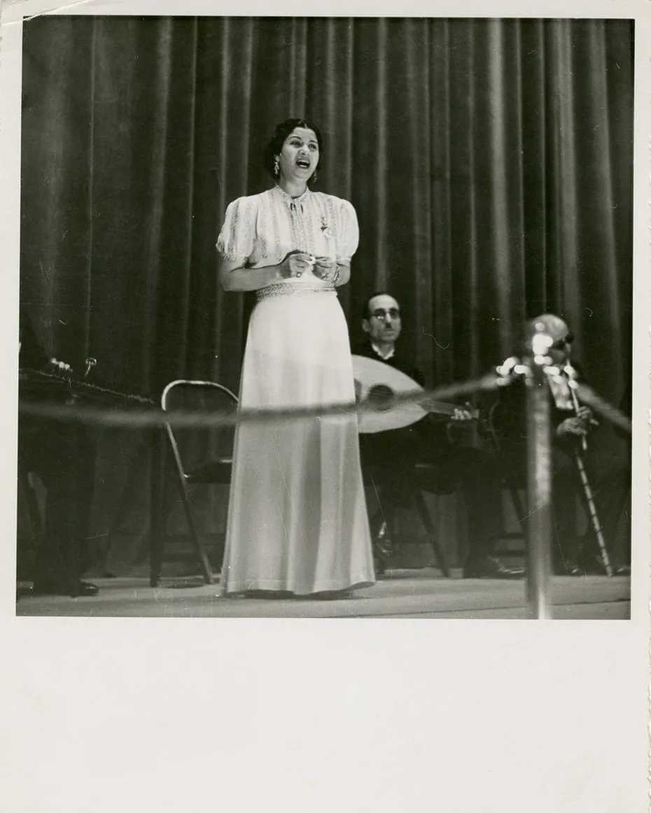 صورة مسرحية بالأبيض والأسود لأم كلثوم تغني بثوب أبيض طويل أمام ستارة مسدلة، ويجلس خلفها عازفا العود والكمان.