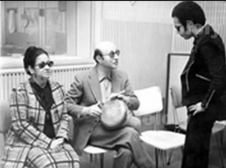 صورة بالأبيض والأسود من الكواليس لأم كلثوم بنظارة شمسية تجلس بجانب الملحن محمد عبد الوهاب الممسك بدف، ويقف شاب.