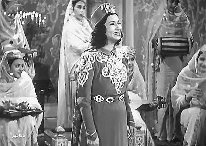 لقطة فيلم بالأبيض والأسود لأم كلثوم تغني مبتسمة بثوب مطرز مزخرف وسط خادمات جالسات في مكان يشبه القصر.