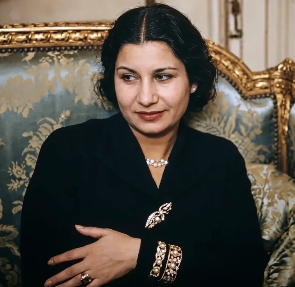 صورة ملونة لأم كلثوم بفستان أسود مع عقد لؤلؤي ودبوس مرصع، تجلس على أريكة مذهبة من البروكار وتنظر جانباً.
