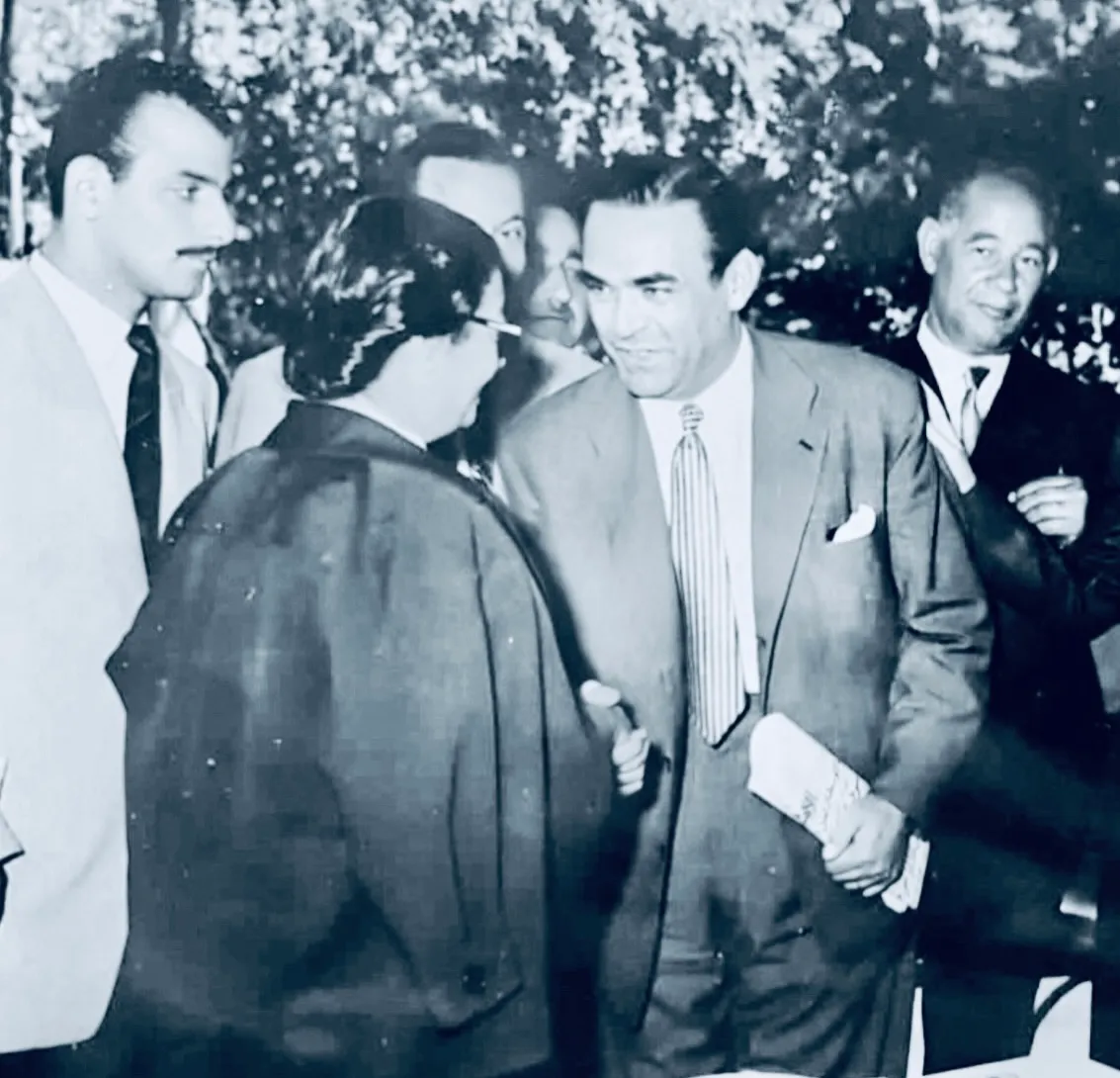 صورة بالأبيض والأسود لأم كلثوم من الخلف بعباءة داكنة تتحدث مع محمود ذو الفقار المبتسم بالبدلة وسط رجال آخرين في الهواء الطلق.