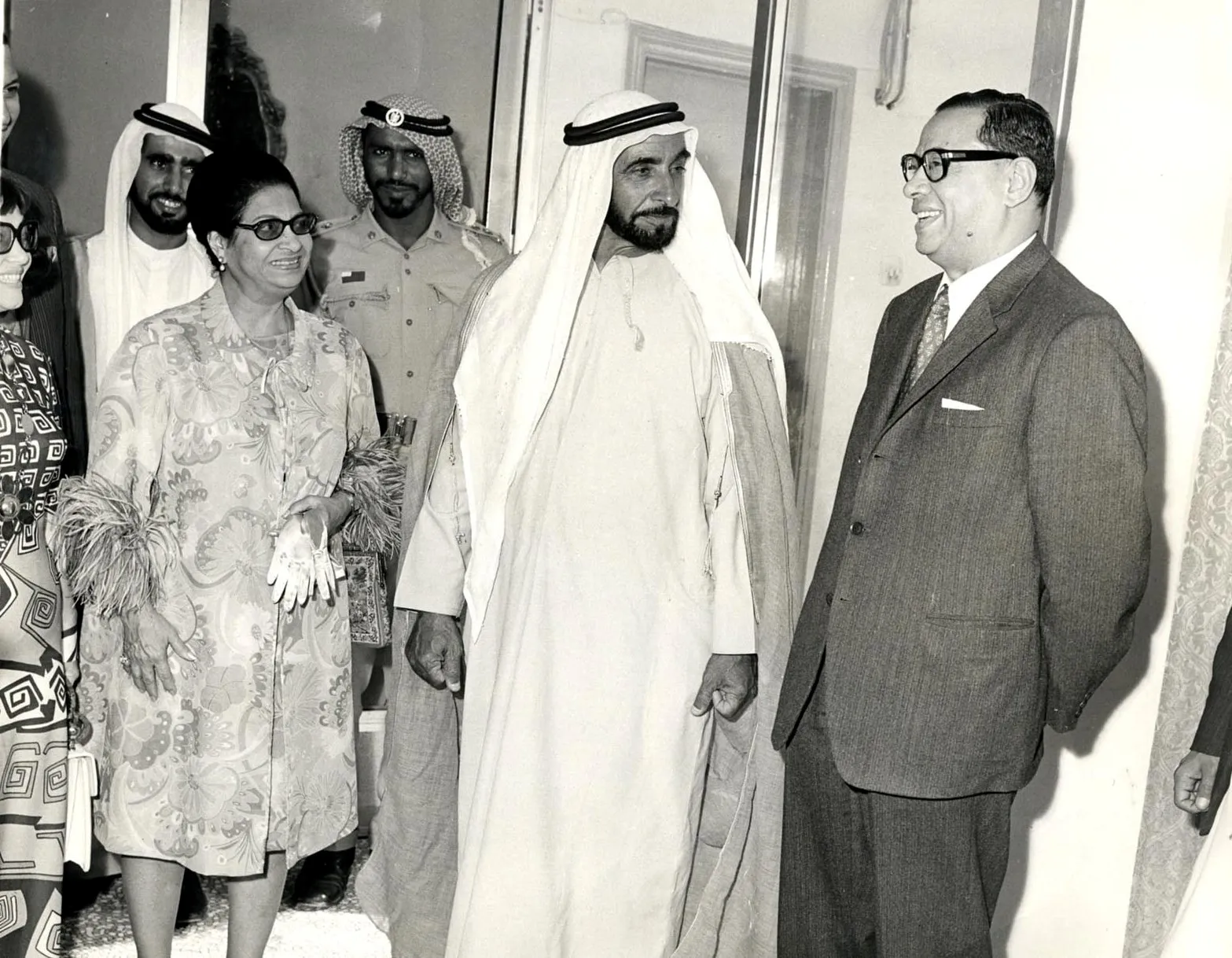 صورة بالأبيض والأسود لأم كلثوم بفستان مزهر ونظارة شمسية تقف بجانب الشيخ زايد بالثوب الأبيض ورجال آخرين.